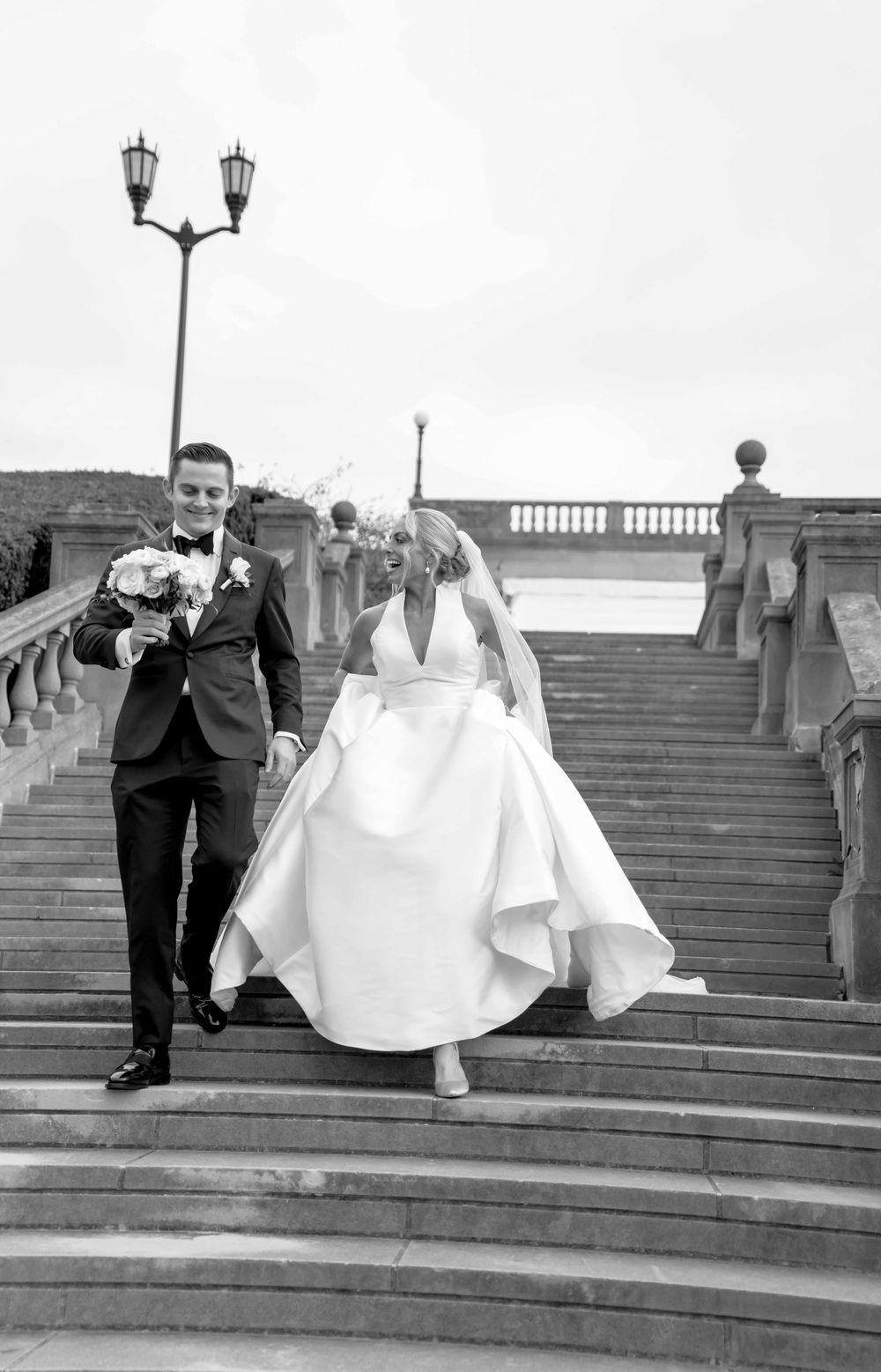 A couple descends stone steps outdoors in formal wedding attire, captured in black and white photography.