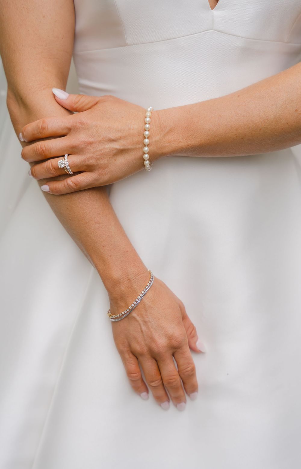 Close-up of hands wearing elegant pearl bracelets and wedding rings against a white dress.