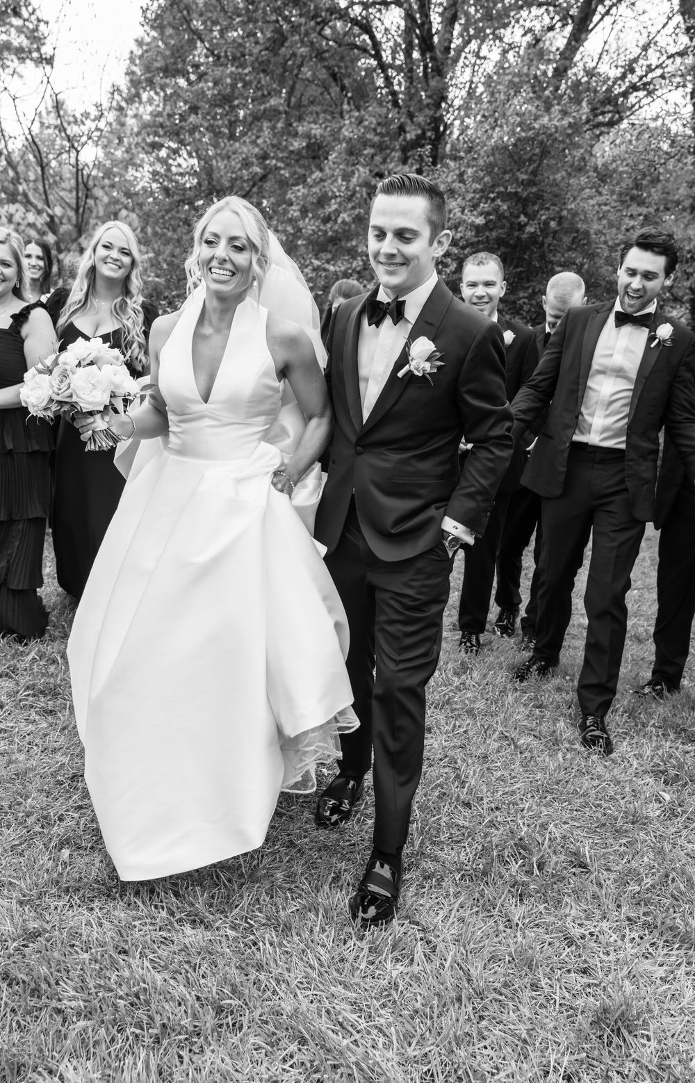 Wedding party laughs and poses together outdoors with bridesmaids in black and groomsmen in suits.