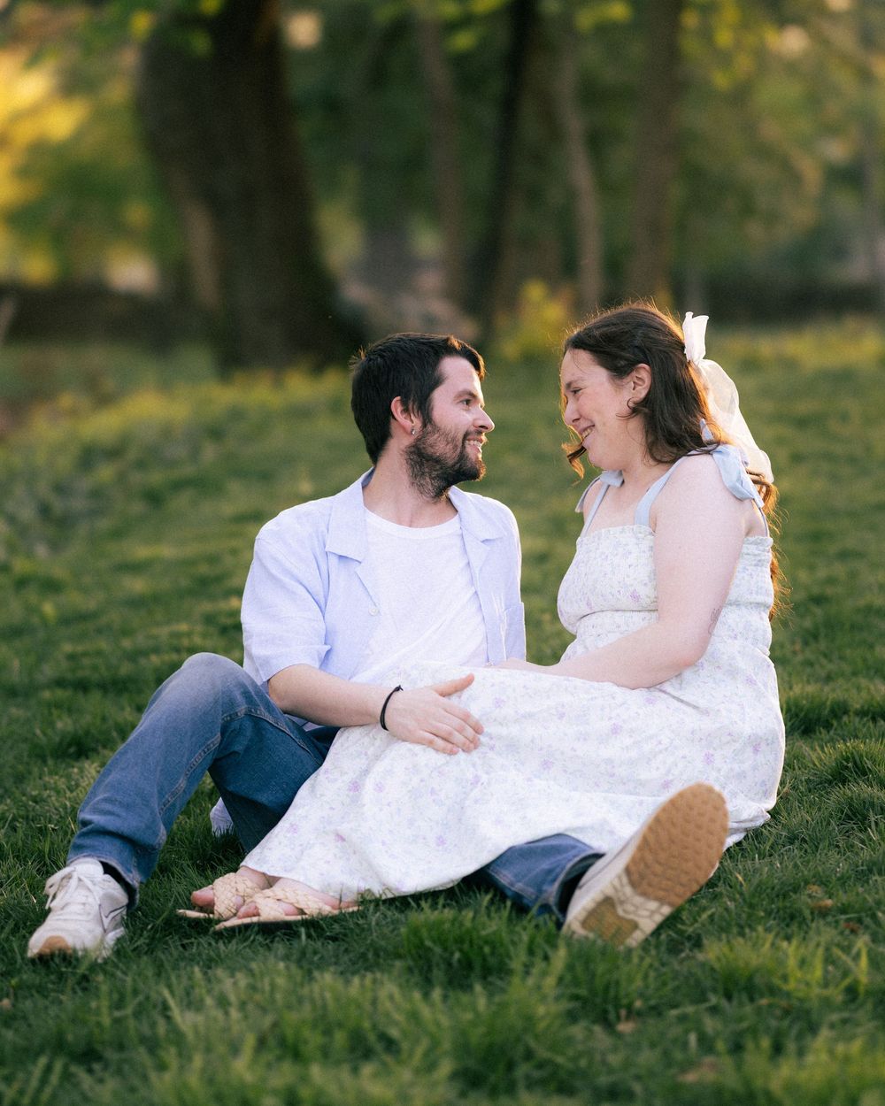 A couple sits on green grass in golden hour light, smiling at each other, woman in floral dress, man in white shirt.