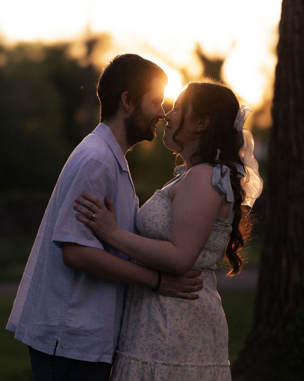 Couple sharing a romantic almost-kiss at golden hour sunset, woman in white dress with veil, man in light blue shirt outdoors.
