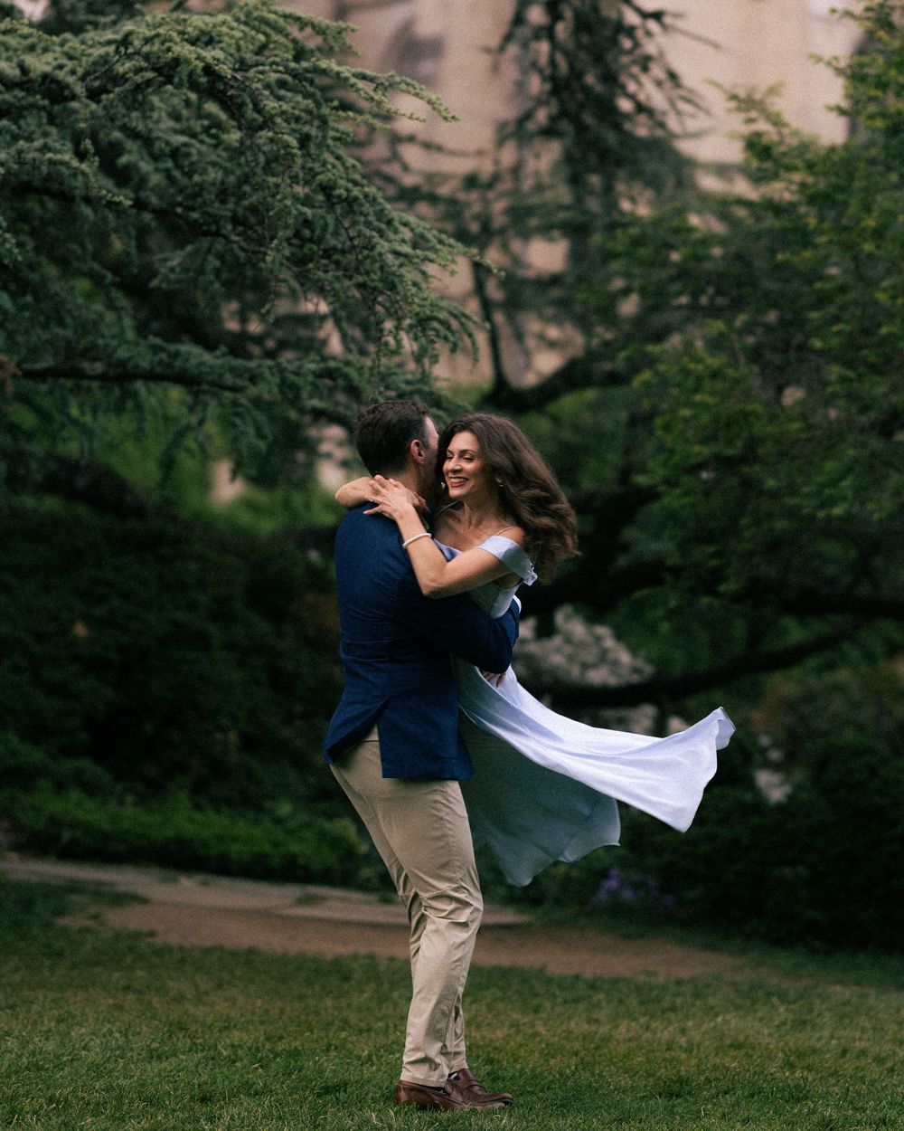 A man in a navy blazer lifts a woman in a white dress outdoors, surrounded by lush green trees.