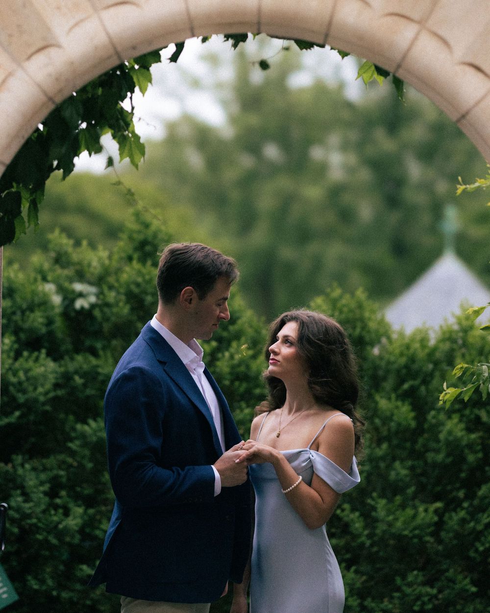 Couple holding hands under a stone arch surrounded by lush greenery, man in navy blazer, woman in off-shoulder gray dress.