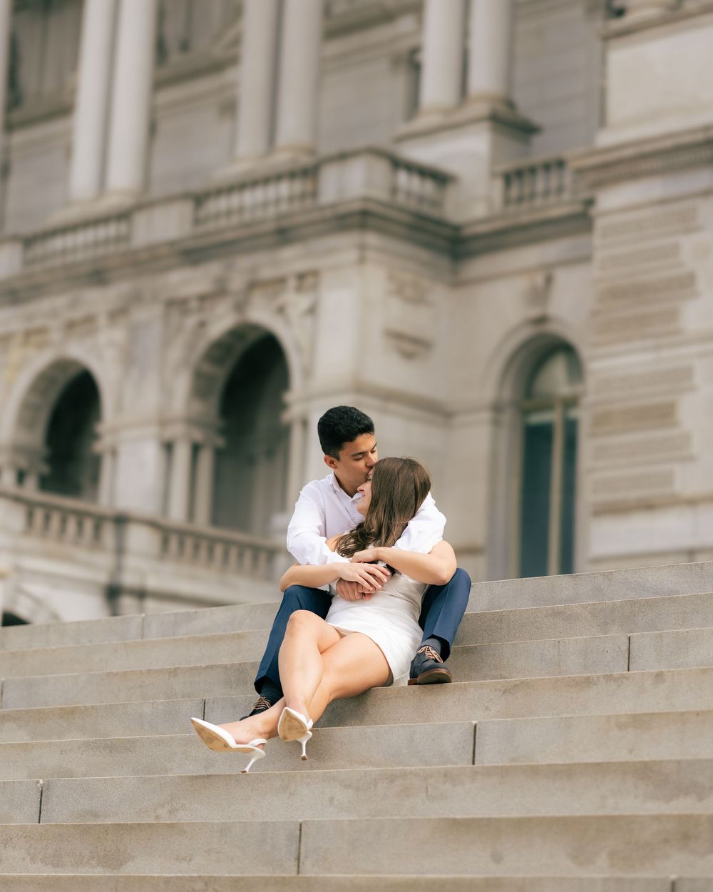 A couple shares an intimate moment sitting on grand stone steps of a historic building with classical architecture.