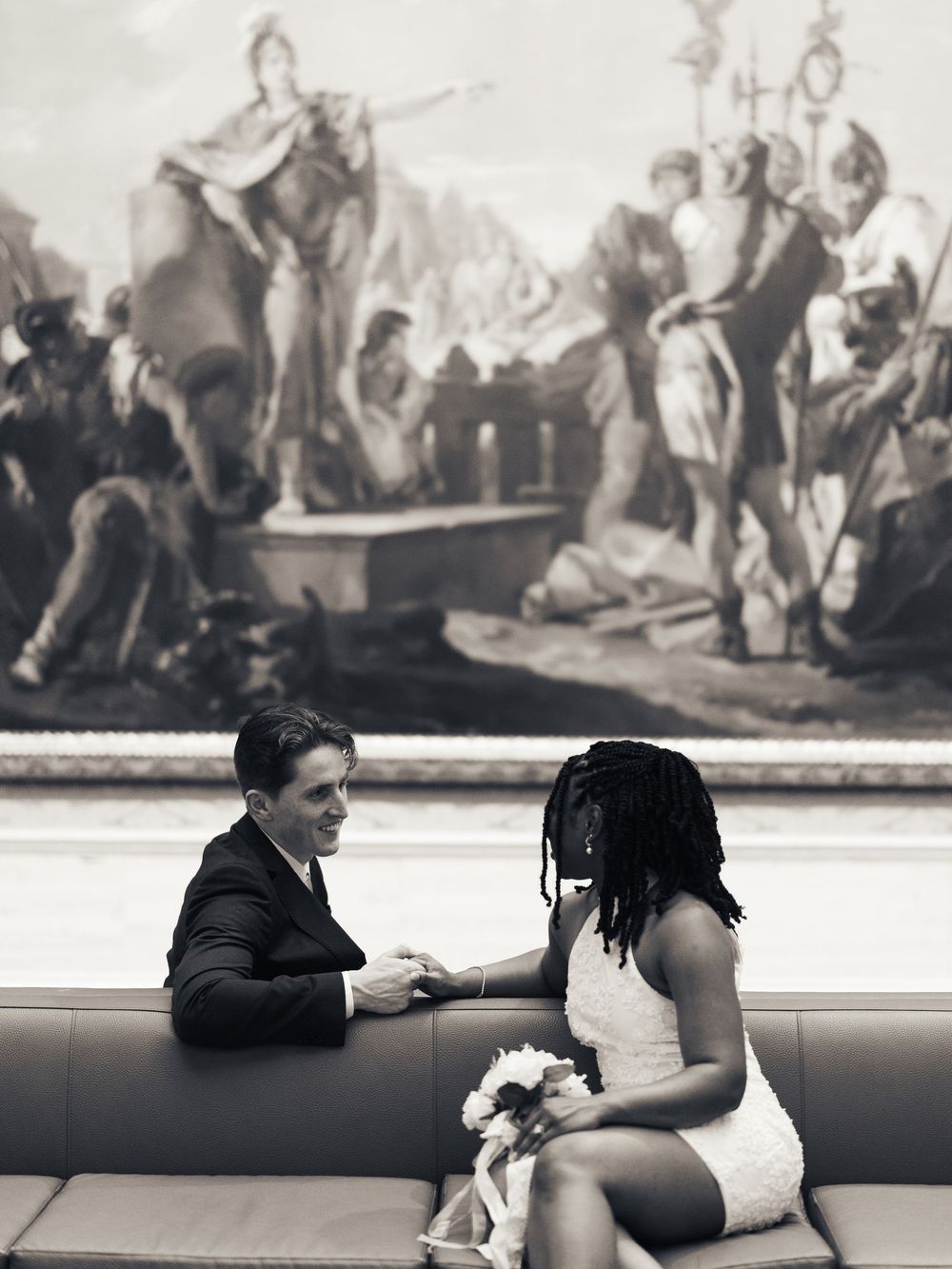 Black and white photo of a couple holding hands on a couch, with a classical painting as backdrop.