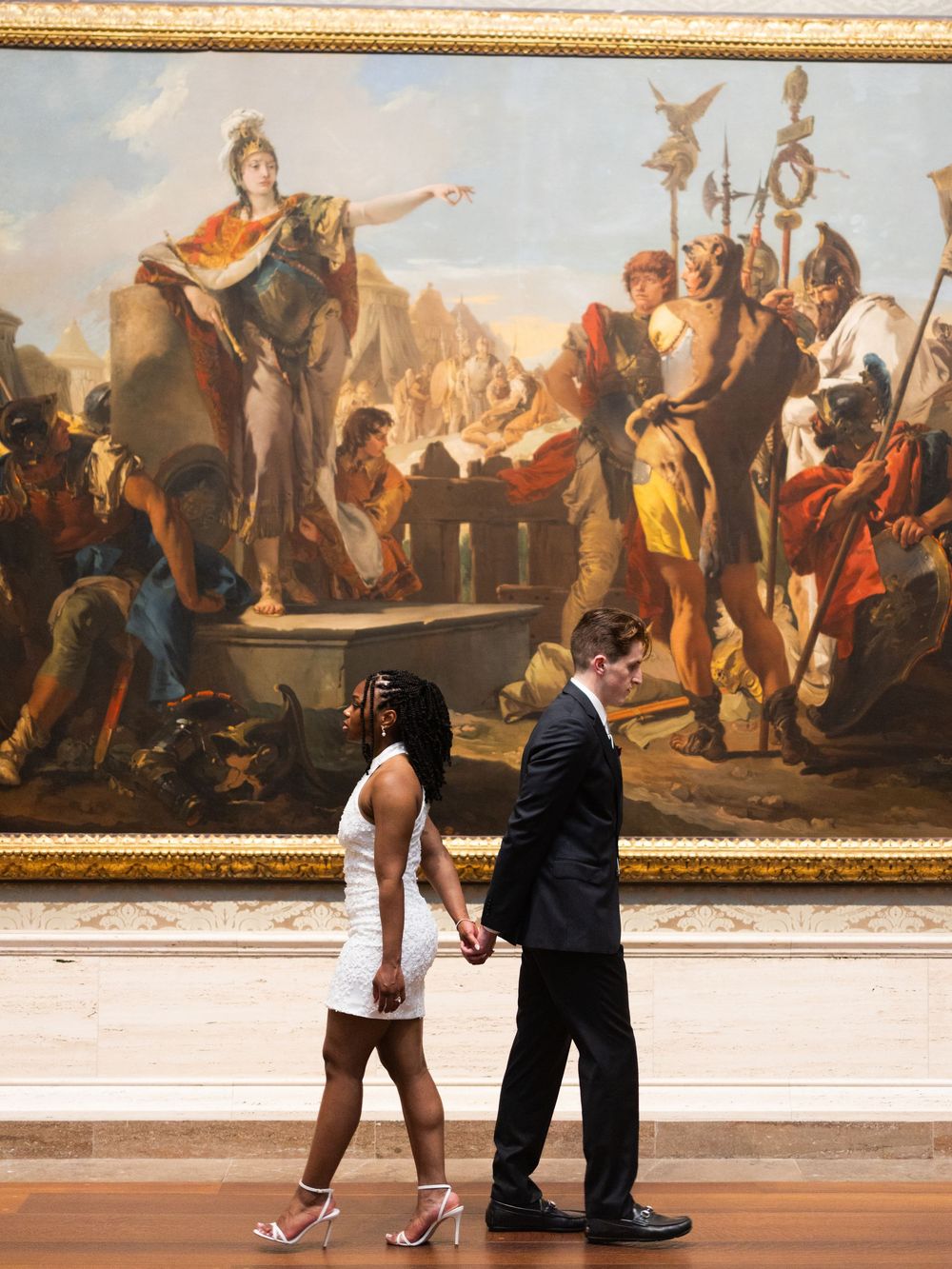 Couple holding hands in white dress and black suit before a grand classical painting in a museum gallery.