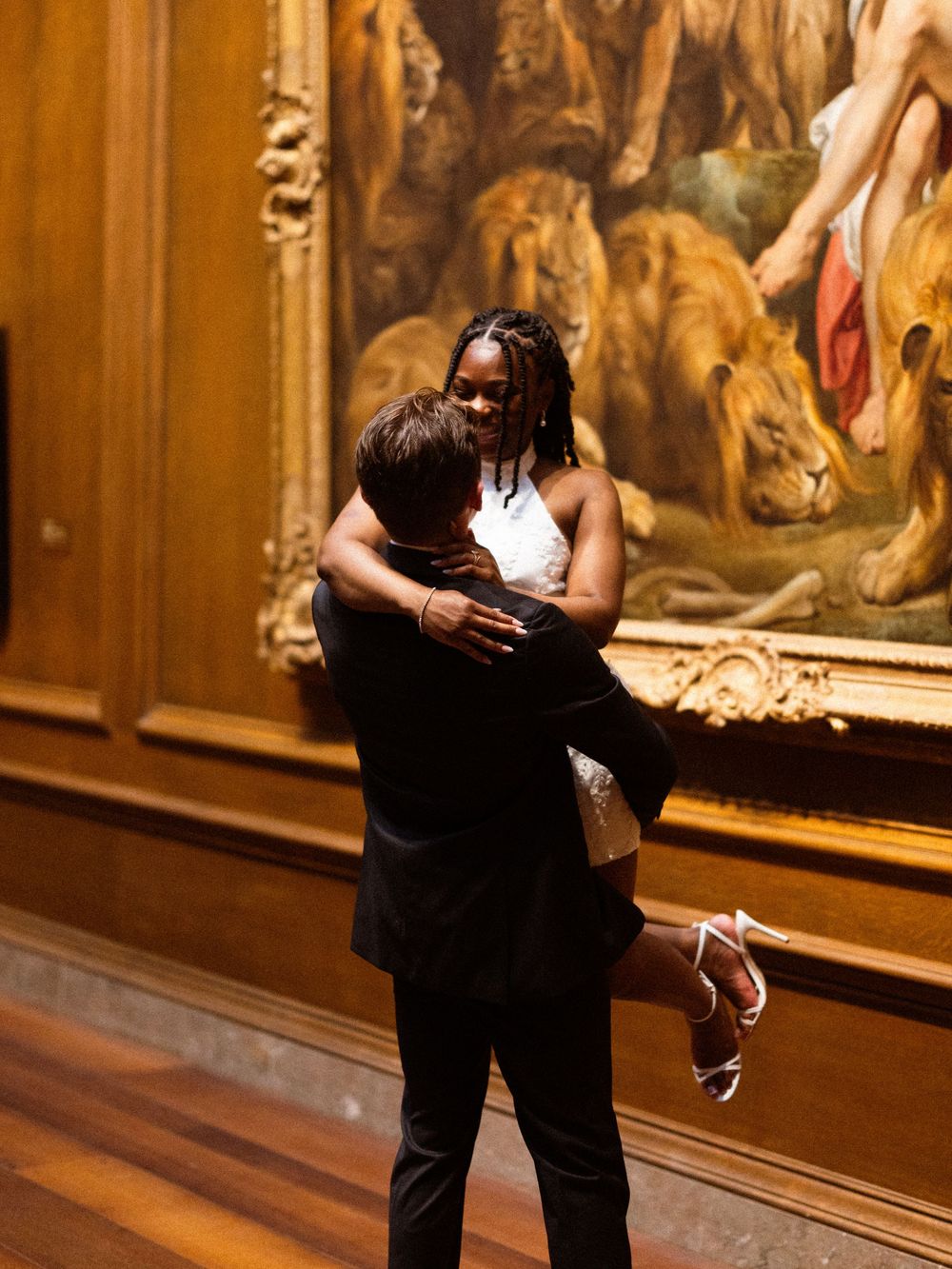 Couple embracing in a museum gallery with a classical painting featuring lions in the background.