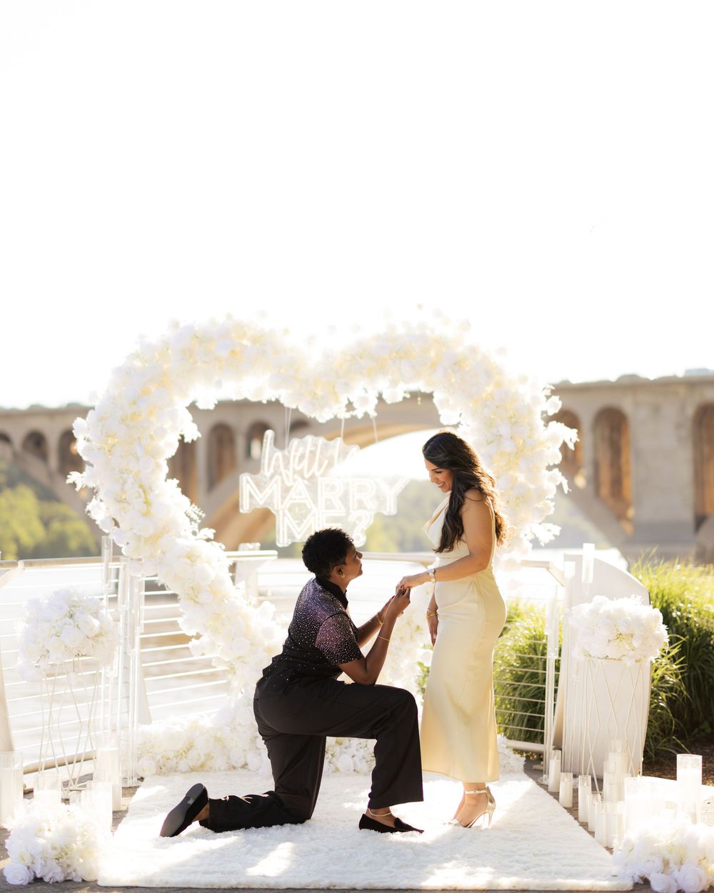 Man proposing to woman under a white floral heart arch with "Will You Marry Me" sign outdoors.