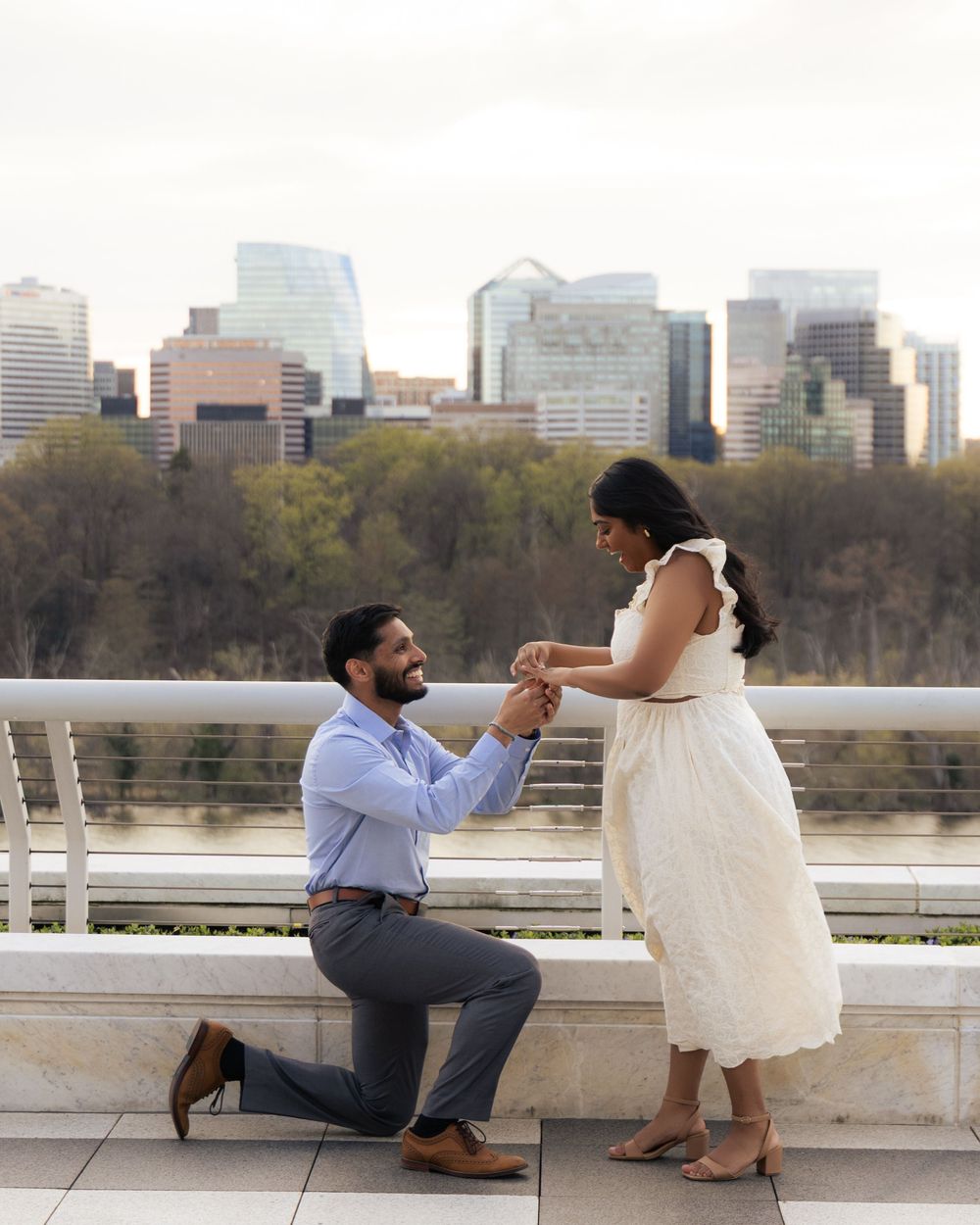 Man proposing on one knee to woman on a terrace with city skyline in the background during engagement shoot.