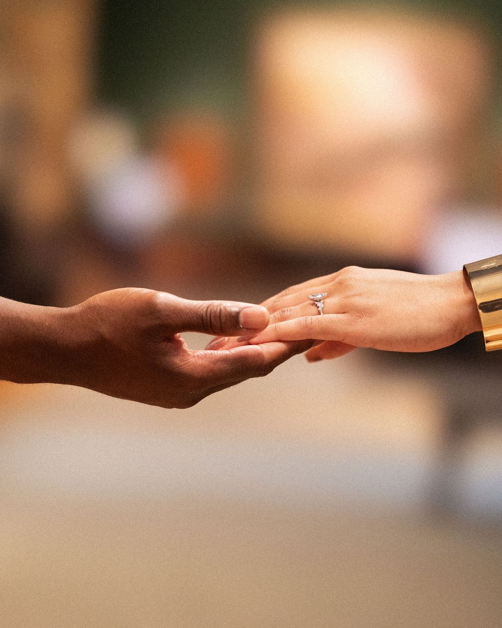 Two hands gently touching fingertips, one wearing a diamond ring and gold cuff bracelet, against a softly blurred background.