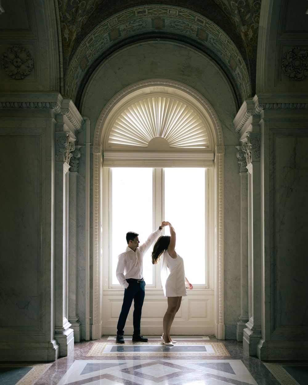 A couple stands in silhouette against a bright arched window in an ornate marble hallway of a historic building.