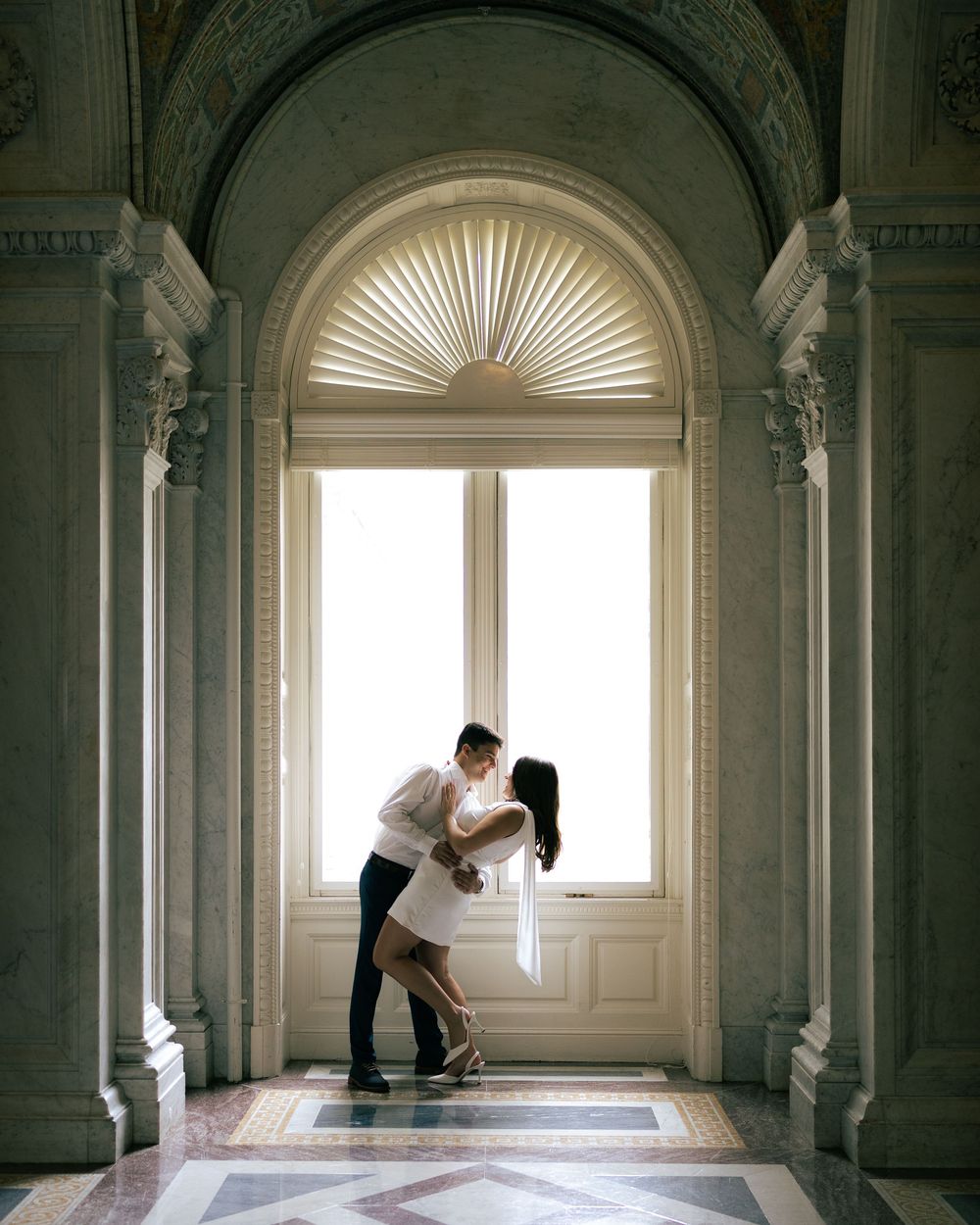 A couple performs an elegant dance pose in a bright classical hallway with arched windows and ornate architectural details.