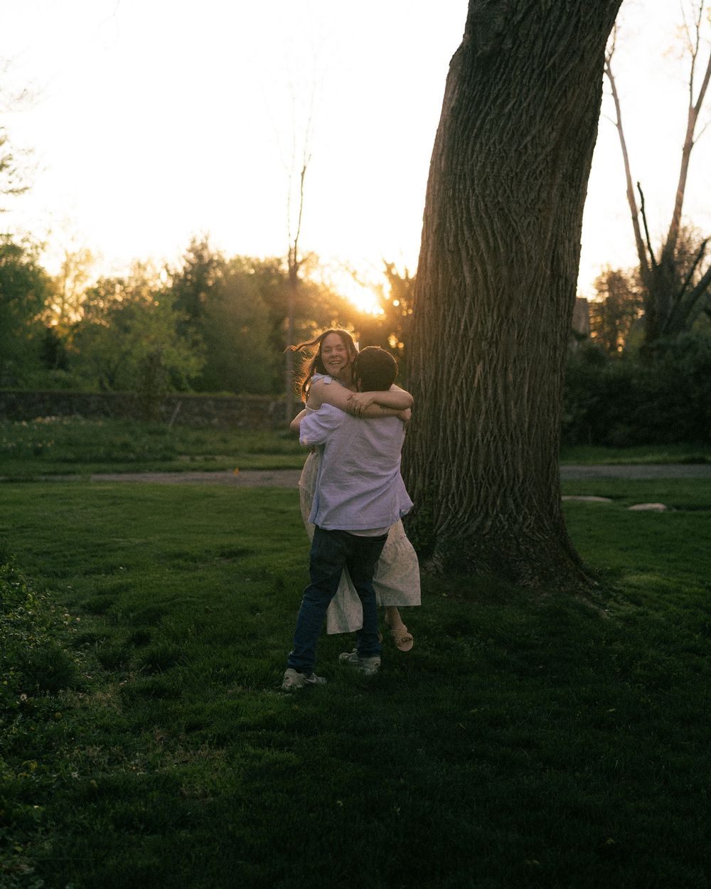 Two people hugging near a large tree in a park at golden hour sunset.