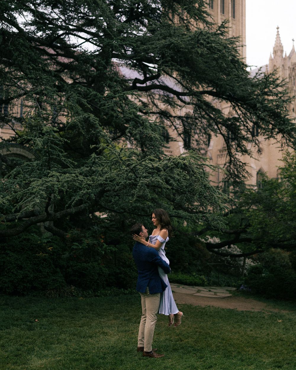 A couple shares a romantic moment as a man lifts a woman in a garden near a historic stone building.
