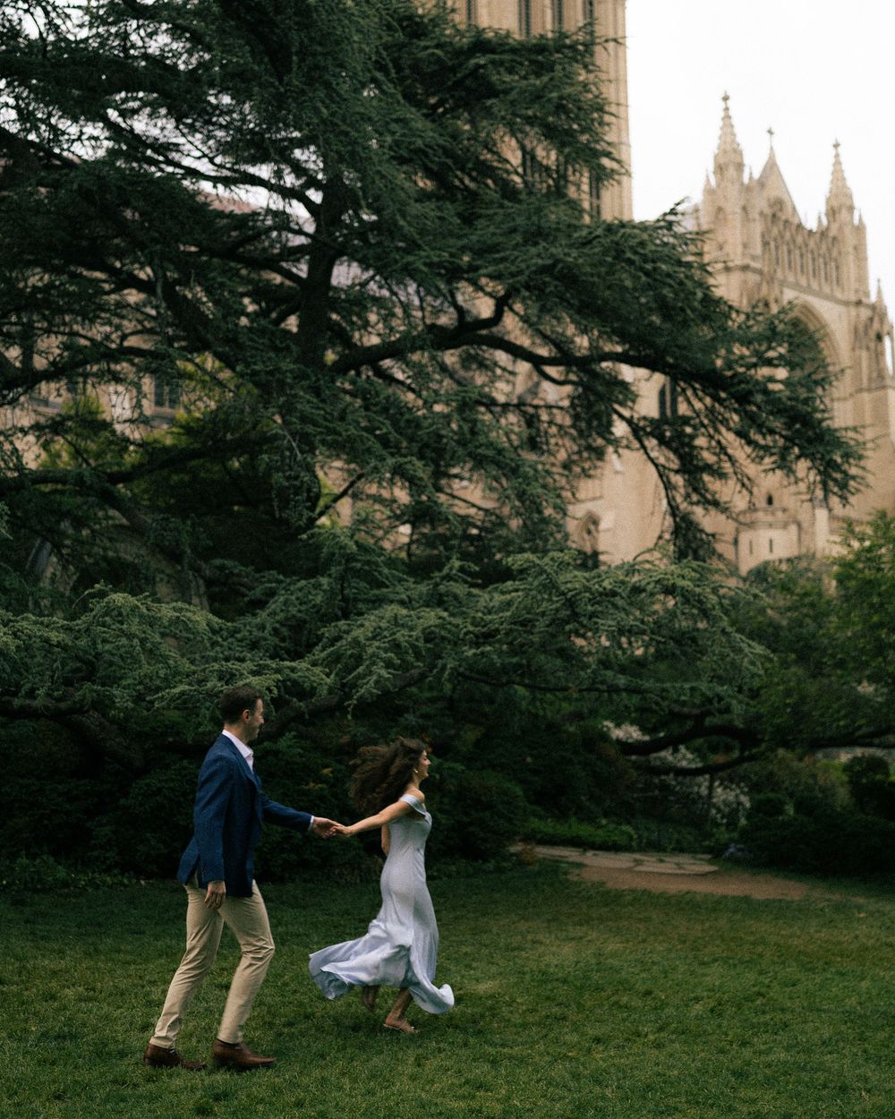 Couple dancing on green lawn before grand cathedral and lush cedar tree in romantic outdoor engagement photo.