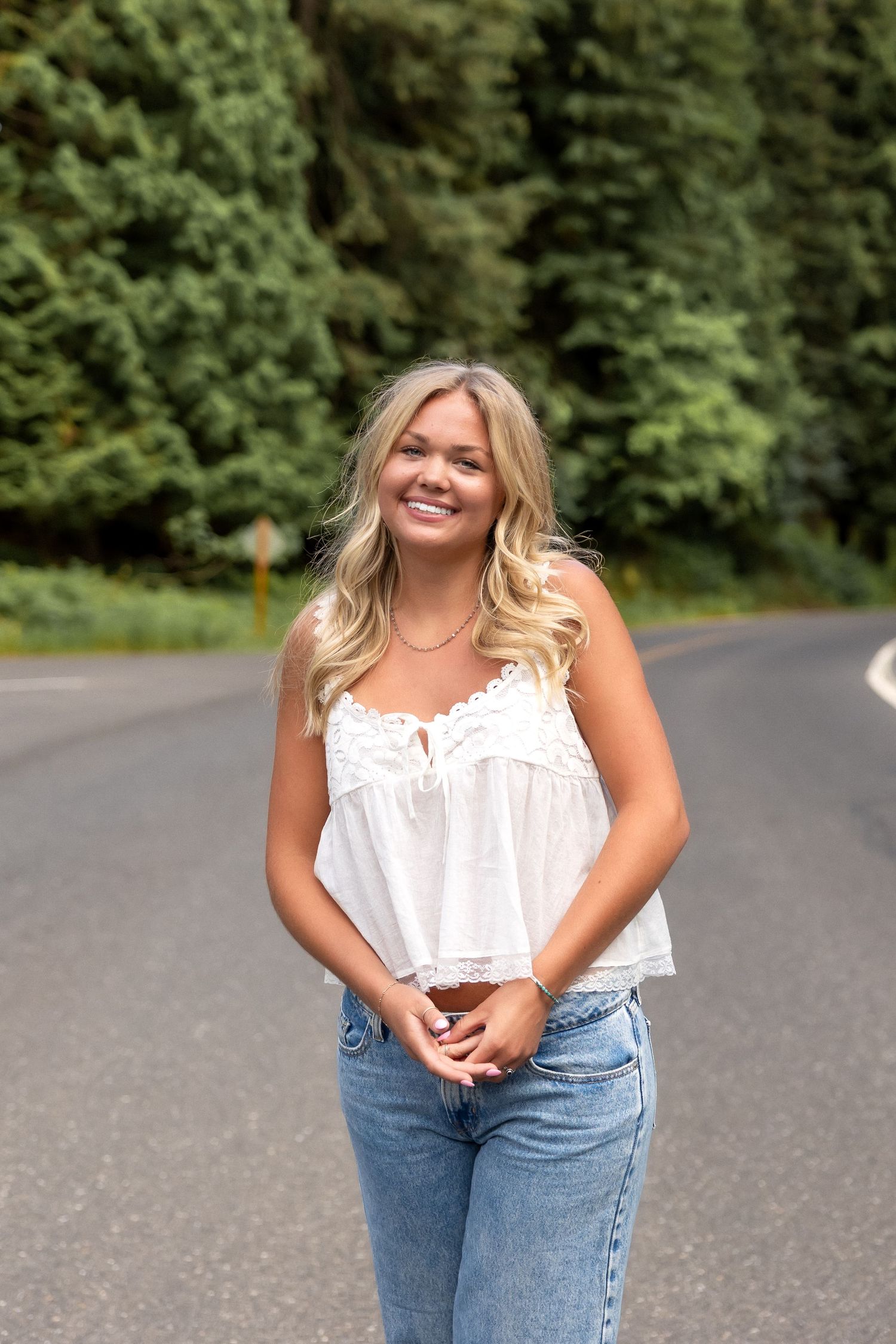 A person in a white sleeveless top and blue jeans poses casually on a tree-lined road.