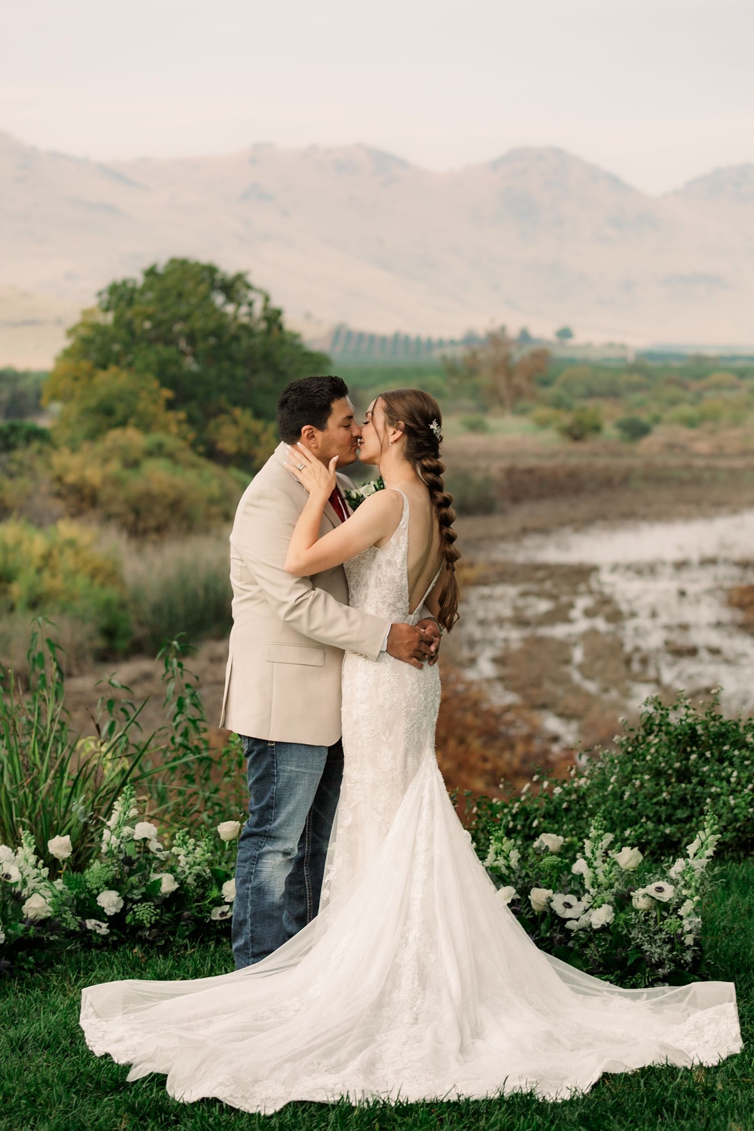 A romantic wedding couple shares a kiss against a backdrop of mountains and wetlands with white flowers in the foreground.