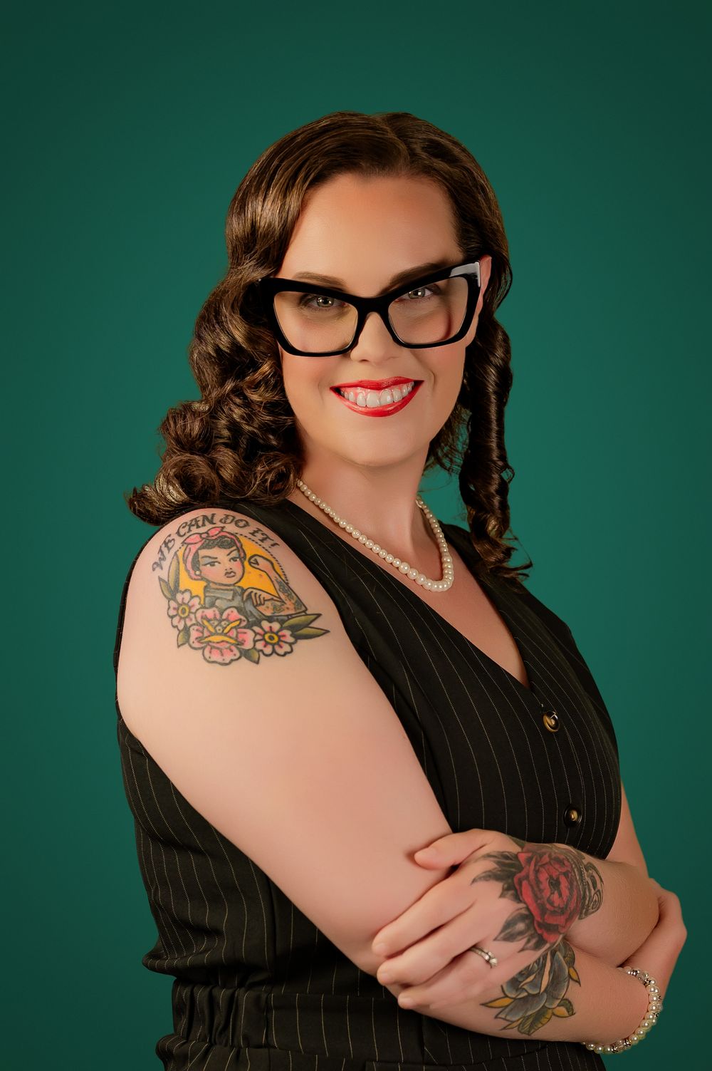 Professional portrait featuring black pinstripe blazer, glasses, and colorful arm tattoos against dark green background.
