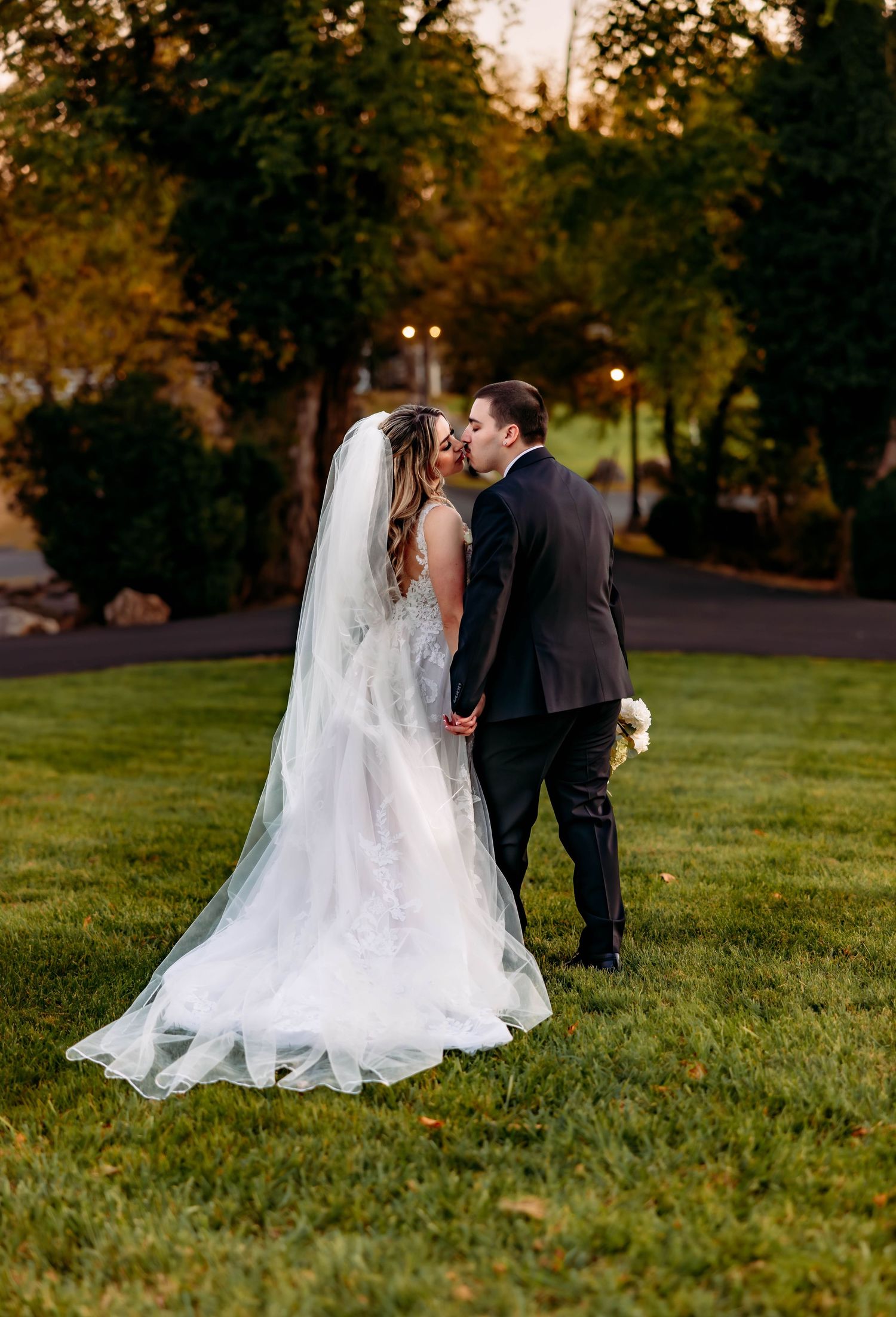 A couple in wedding attire shares a romantic moment on a grassy lawn during golden hour.