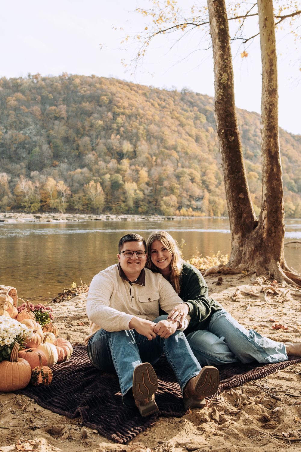 Happy couple sits on a blanket by a river with pumpkins and fall decorations during autumn.