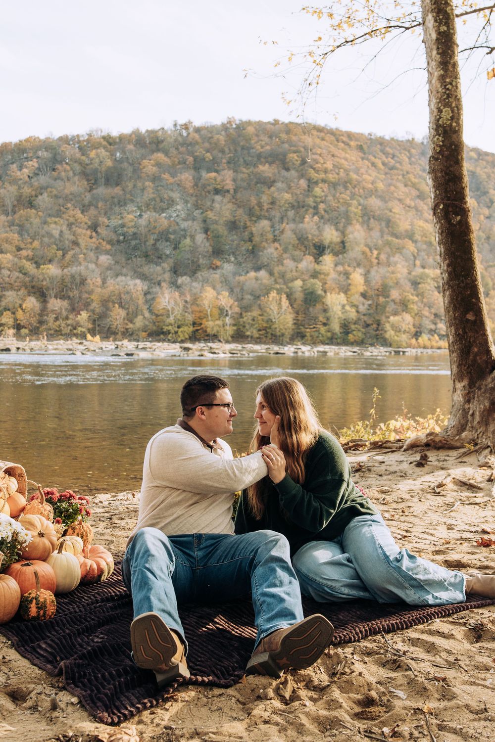 Couple sits on a blanket by a river holding hands surrounded by pumpkins and autumn scenery.