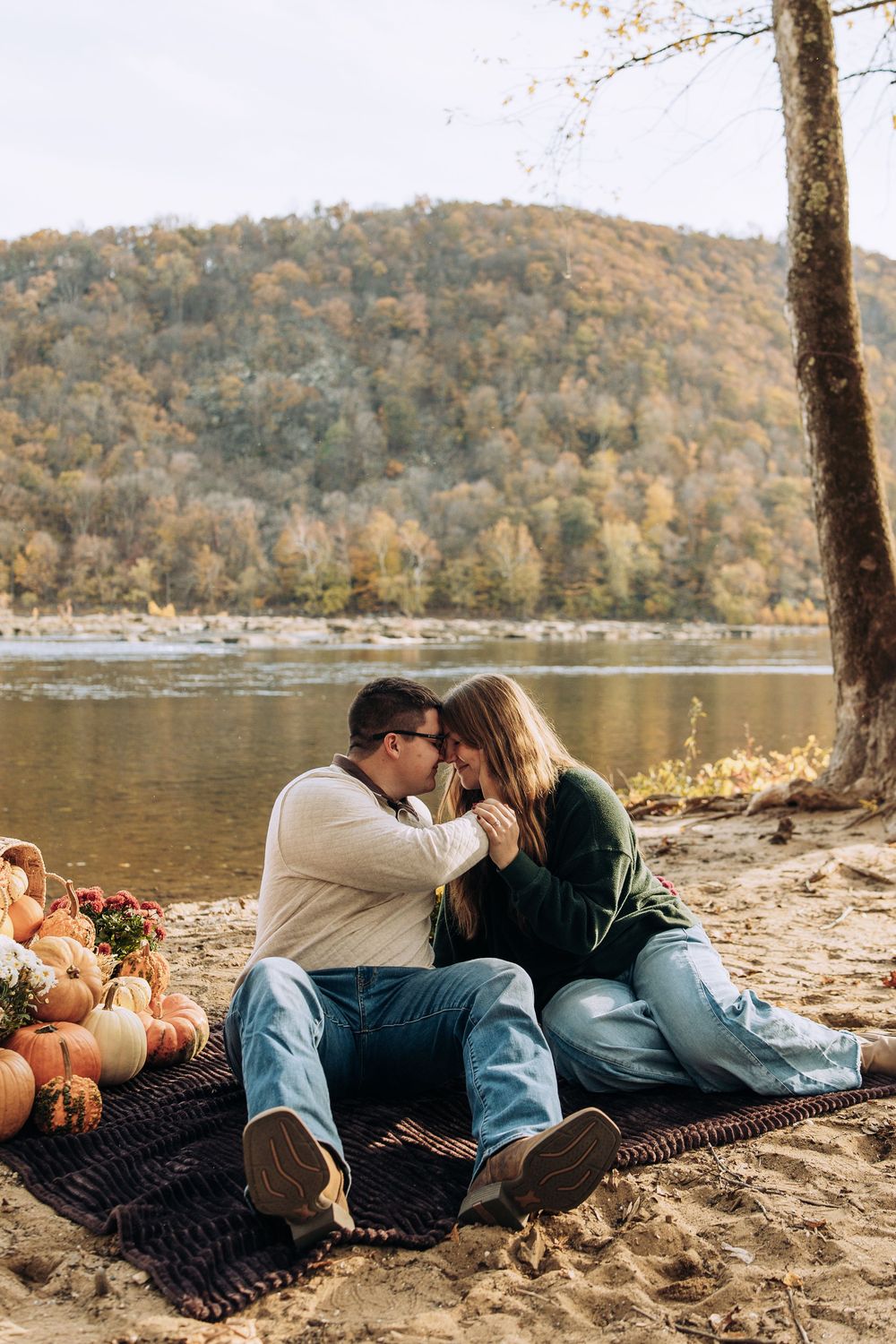 Romantic couple leans in for a kiss while sitting on a blanket by a river with fall pumpkins.