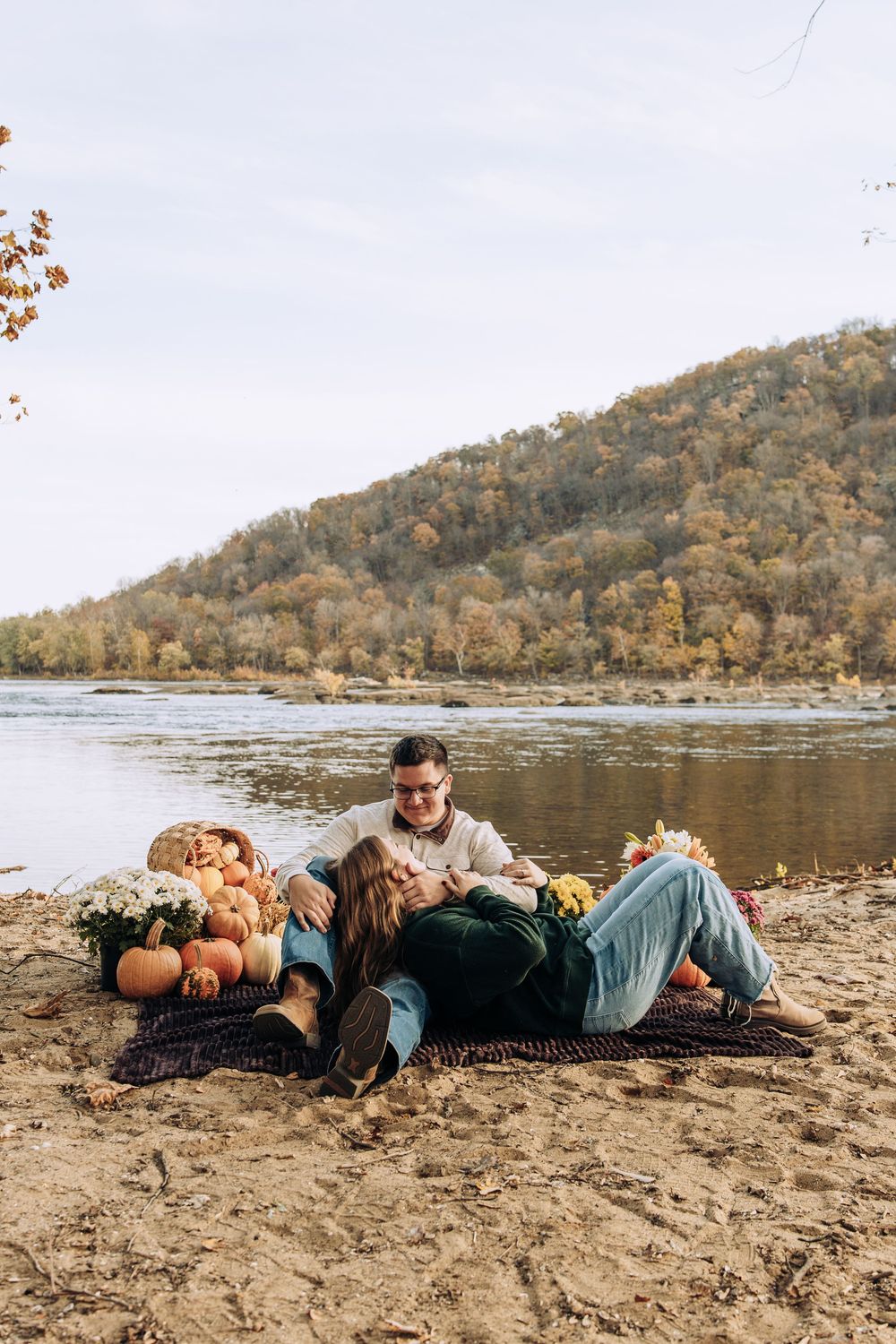 Couple relaxes on a blanket by a river with pumpkins and flowers during a fall picnic.