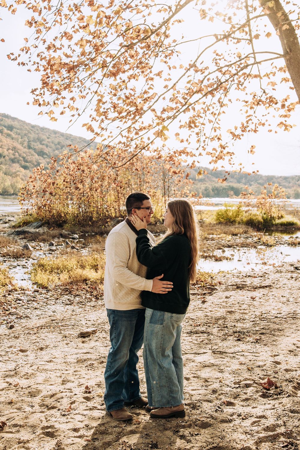 Couple embraces and kisses under autumn trees on a sandy riverbank with colorful fall foliage.