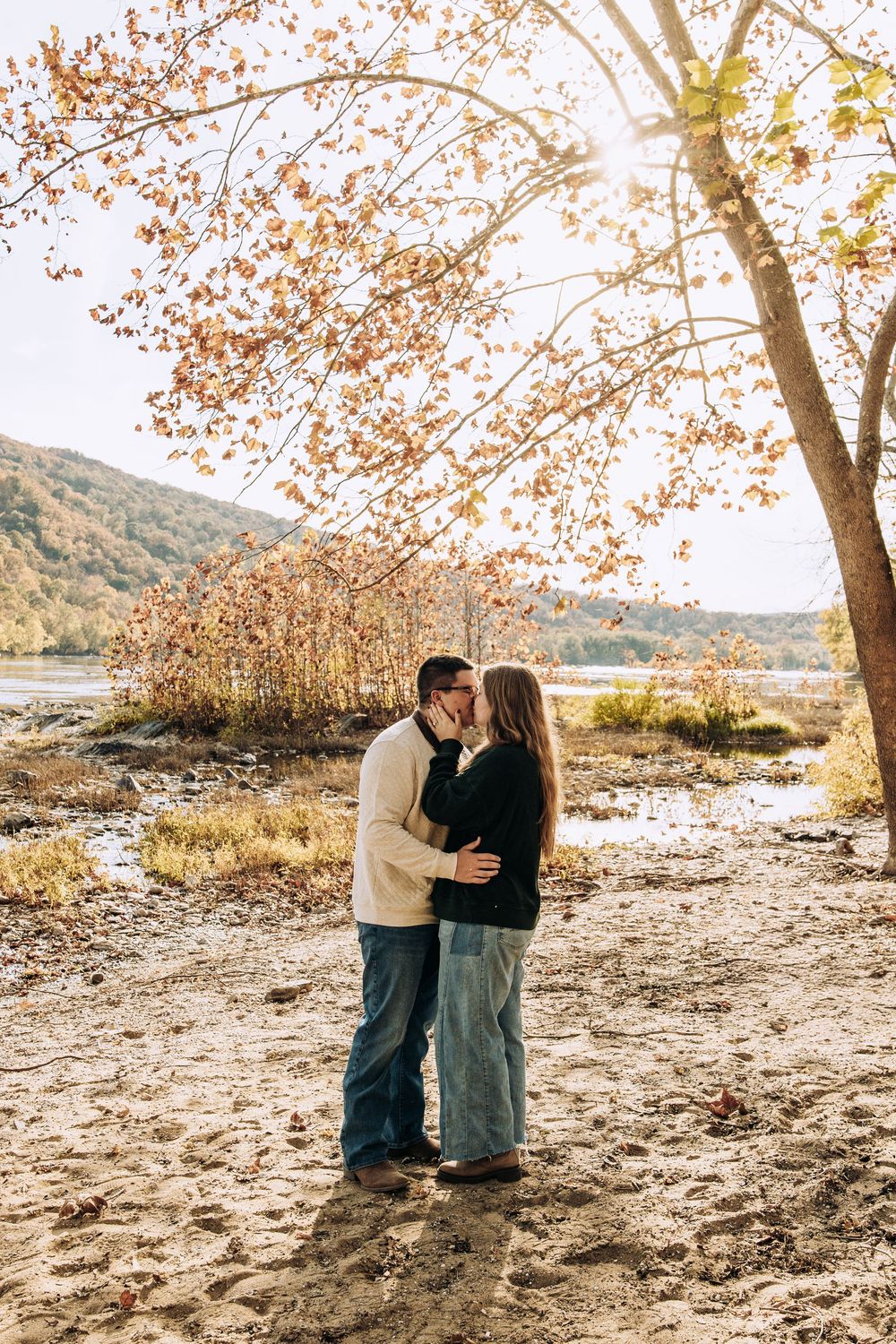 Couple shares a romantic kiss under a sunlit autumn tree on a sandy riverbank.