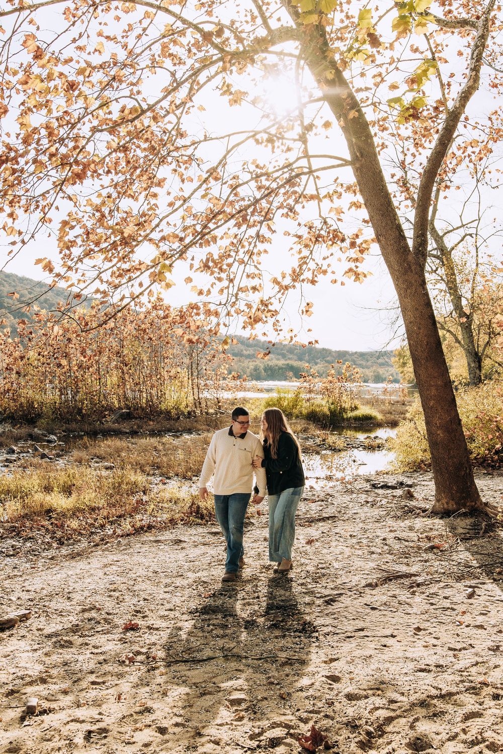 Couple holding hands walking on sandy path surrounded by autumn trees near a lake.