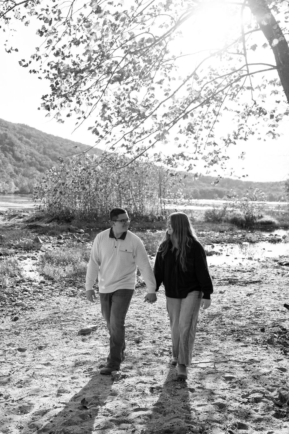 Black and white photo of couple holding hands walking on sandy path with autumn trees.