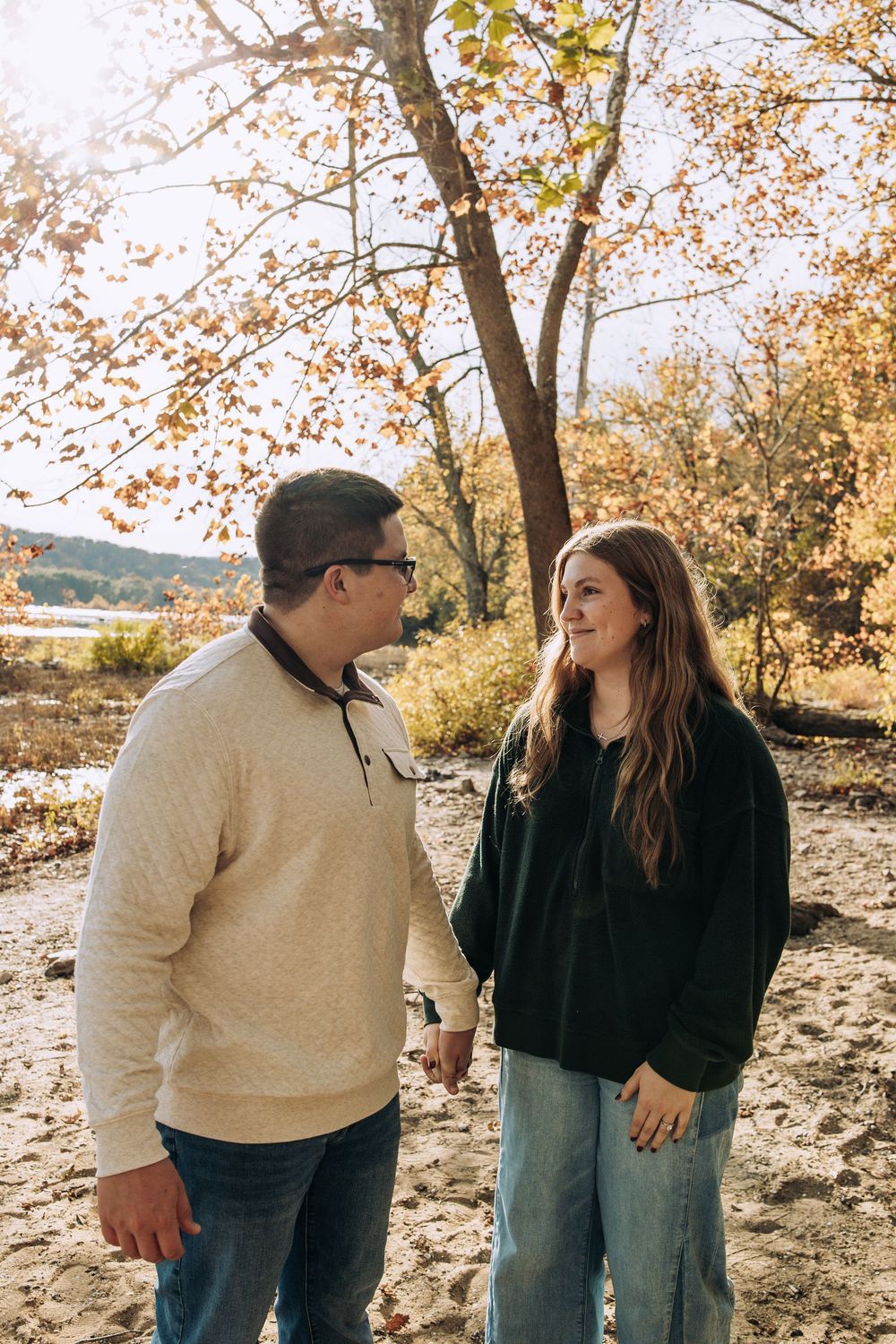 Couple holding hands and smiling at each other on a sandy beach with fall foliage.