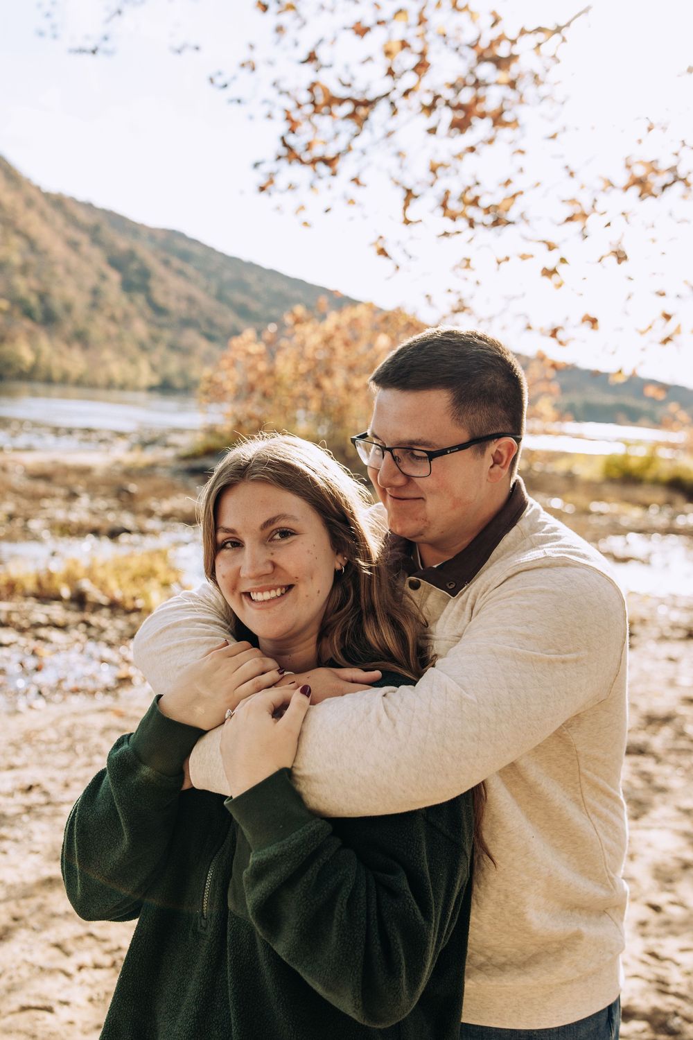 Man embracing woman from behind as they smile outdoors in autumn near a lake.