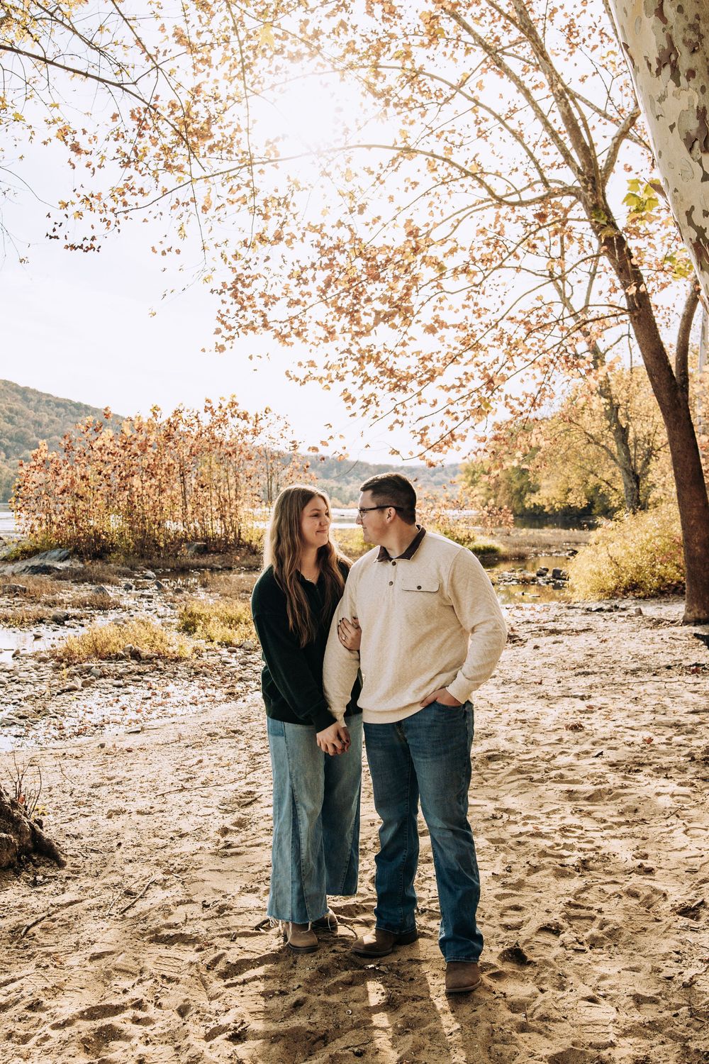 Couple holding hands and gazing at each other under golden autumn trees by a lake.