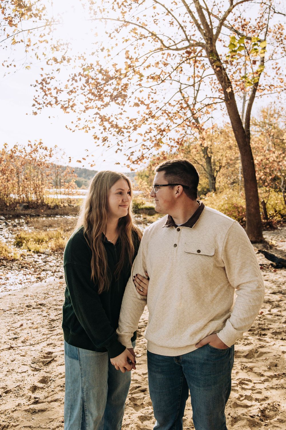 Couple holding hands and looking at each other on a sandy path with fall foliage.