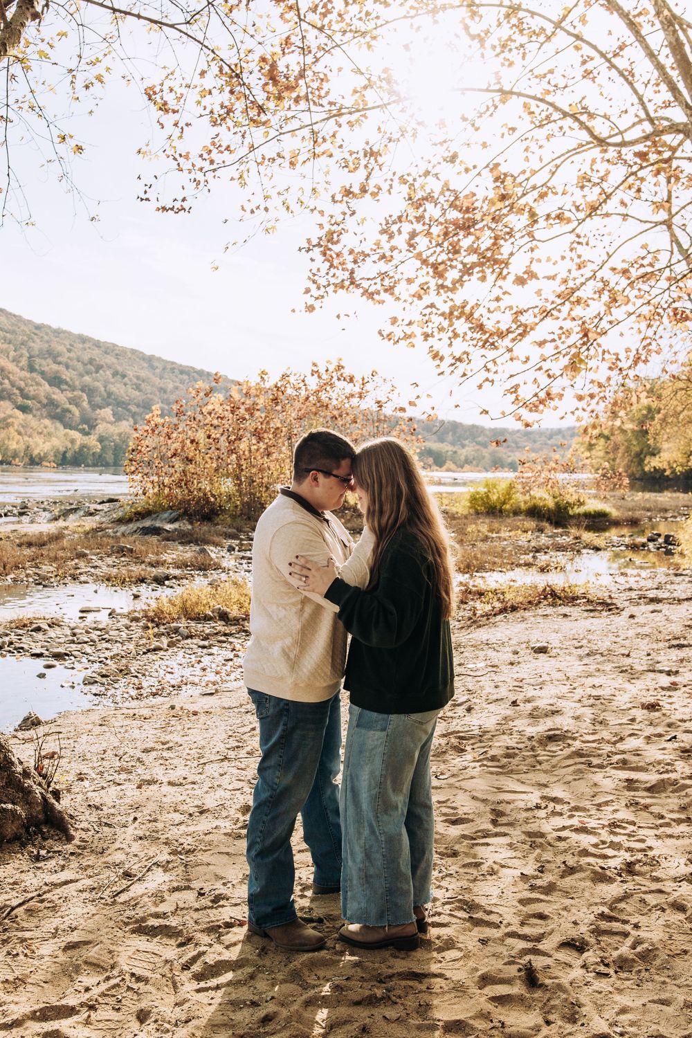Couple standing close together forehead to forehead on sandy beach with autumn trees.