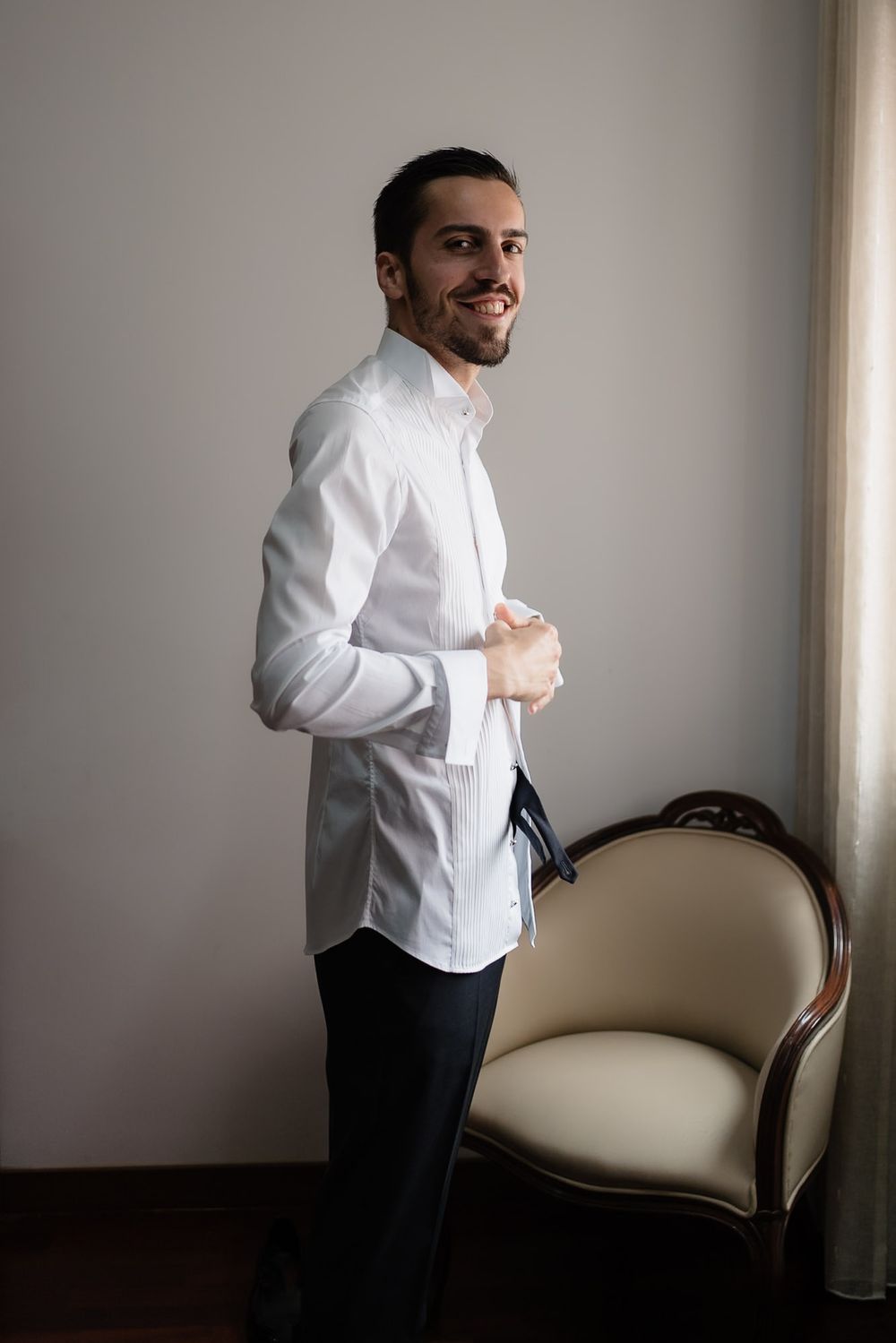 A person in formal attire stands near a cream-colored loveseat in a bright room.