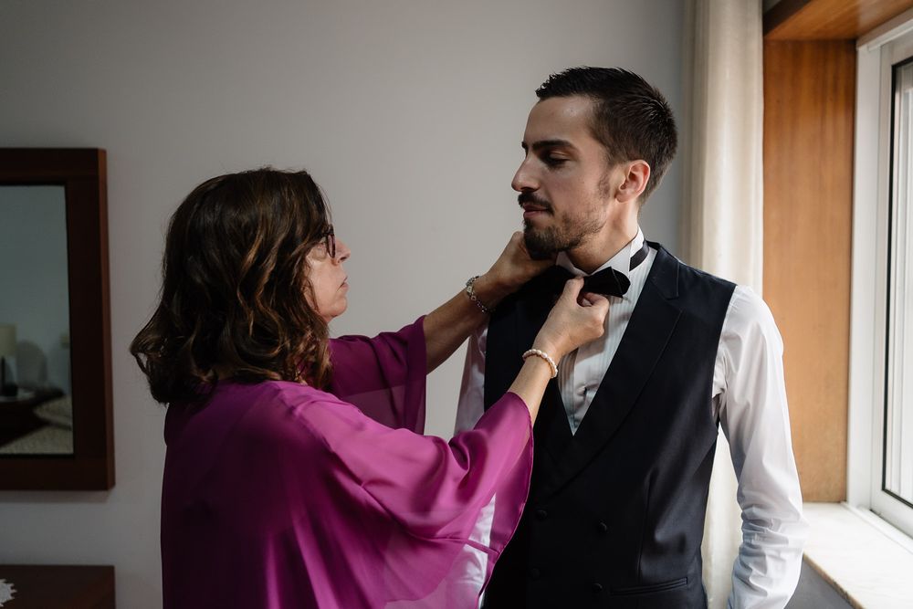 A couple in formal attire share a tender moment as they prepare for a special occasion near a window.