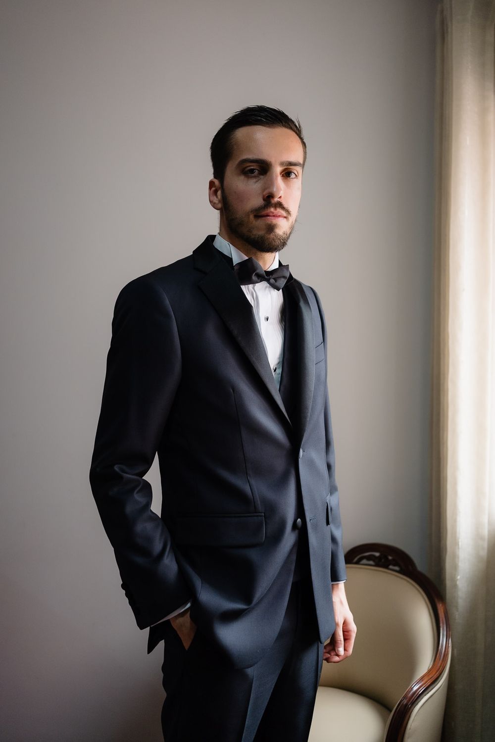 A man in a formal black suit and white shirt stands against a light wall near an elegant vintage chair.