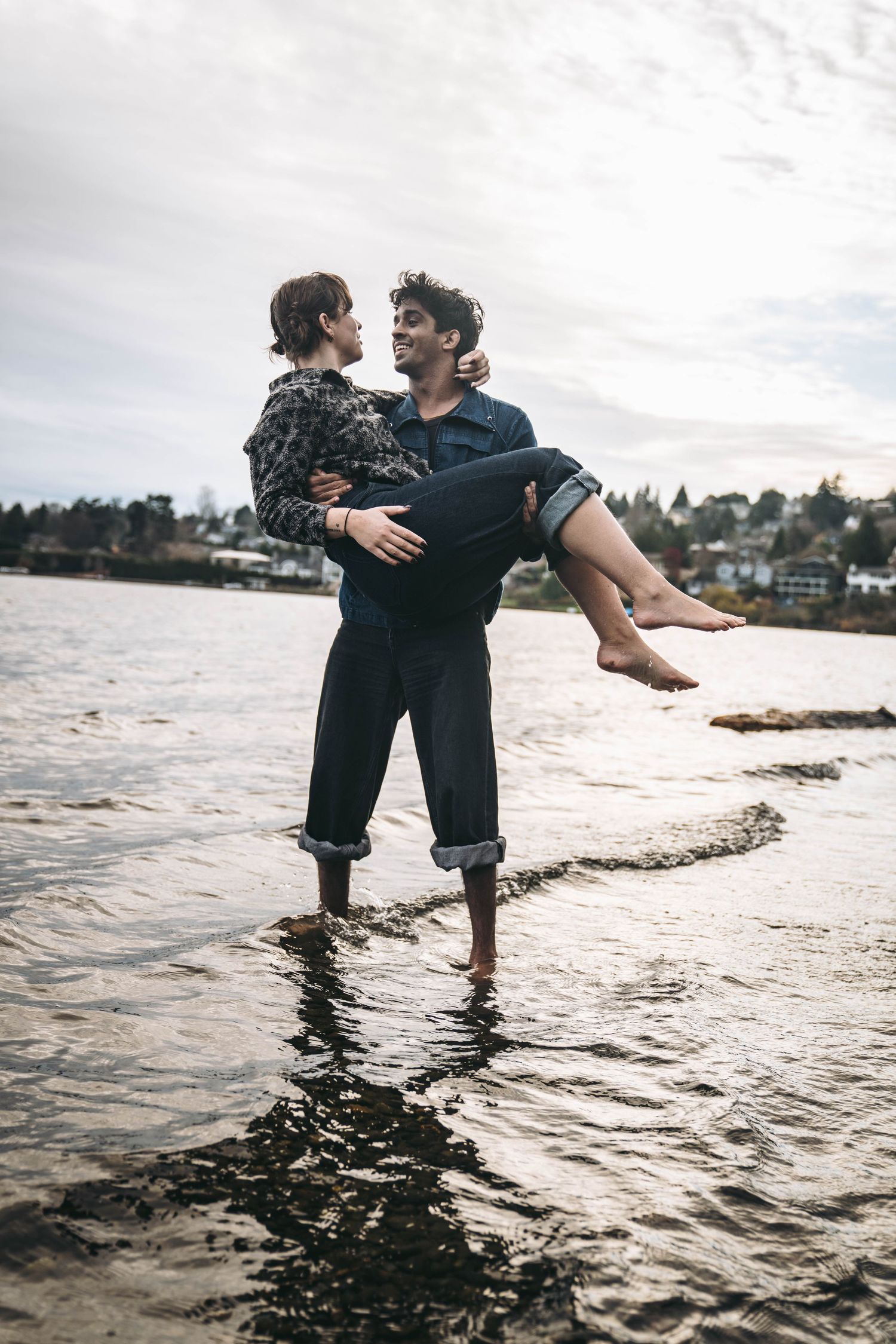 A man carries a woman in his arms while standing in shallow water along a rocky shoreline under a cloudy sky.