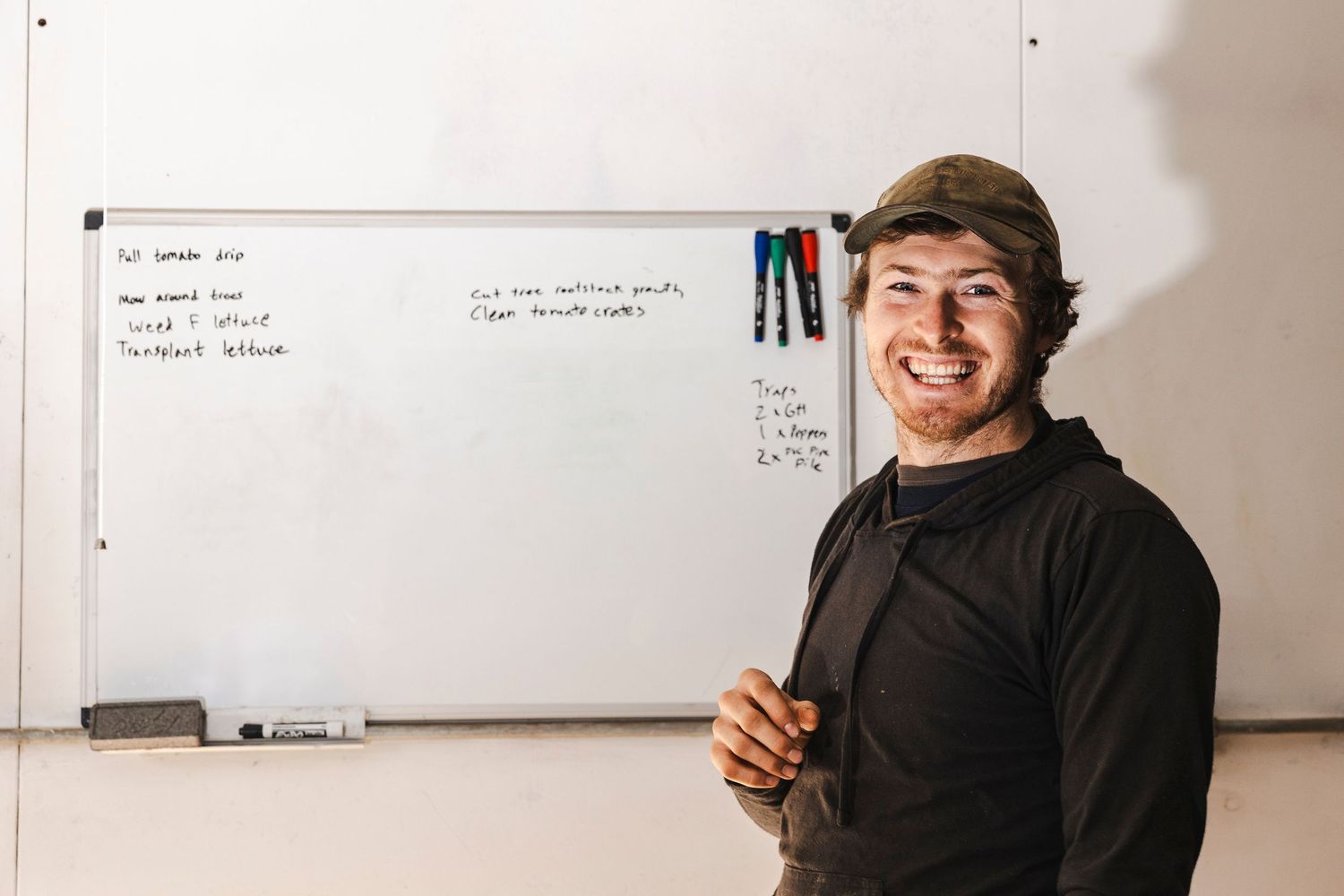 Someone in a black hoodie smiles while standing in front of a whiteboard in a classroom setting.