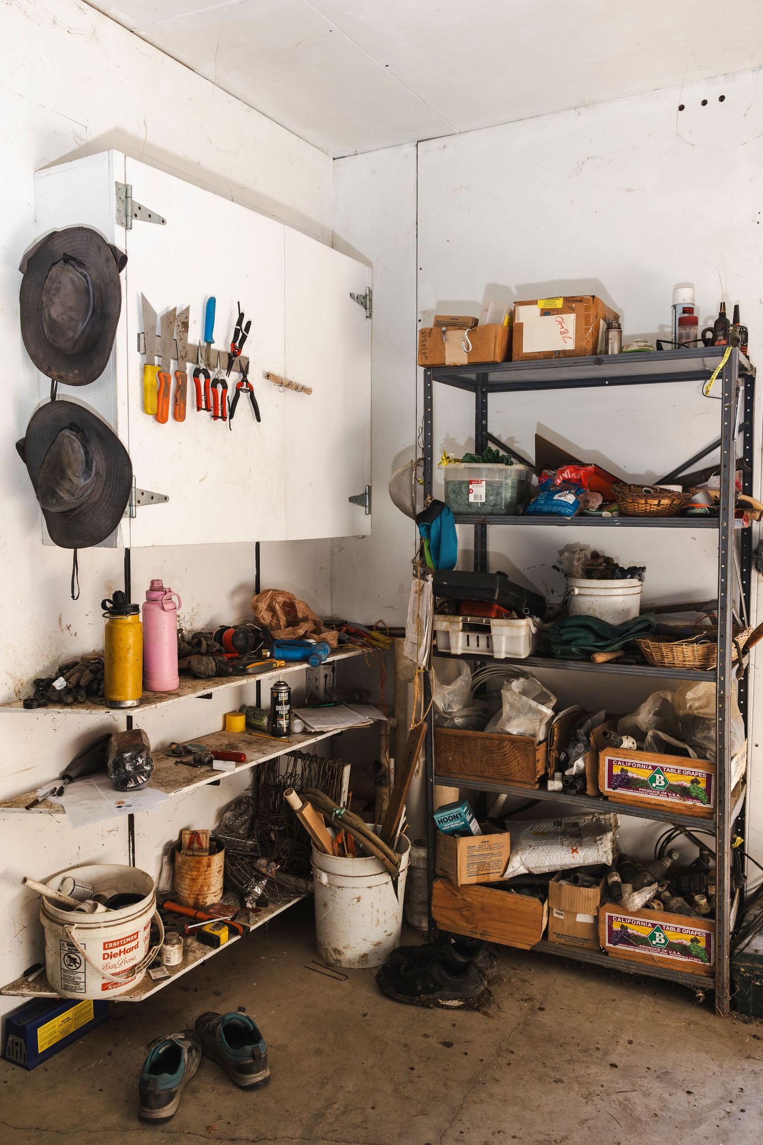 Cluttered garage storage shelves filled with tools, buckets, hats, and miscellaneous household items against white walls.