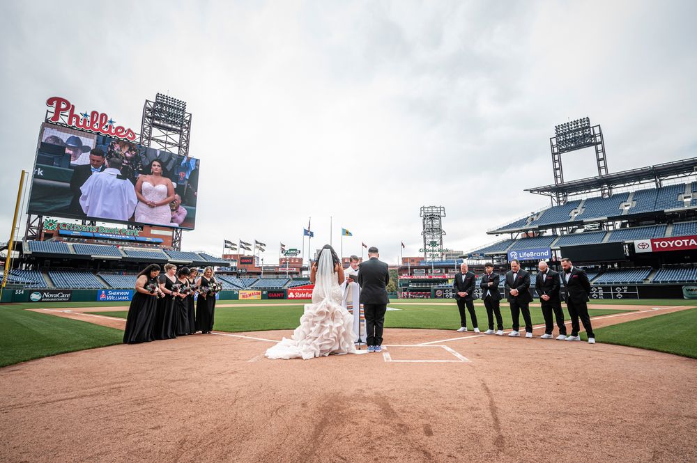 Wedding party poses for photos on a baseball diamond with stadium seating and Phillies scoreboard in background.