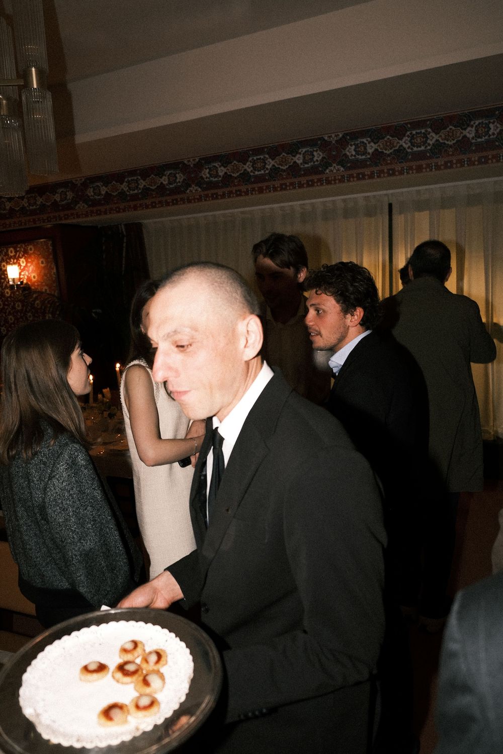 People in formal attire at a dark reception gathering around a white frosted cake with decorative toppings.
