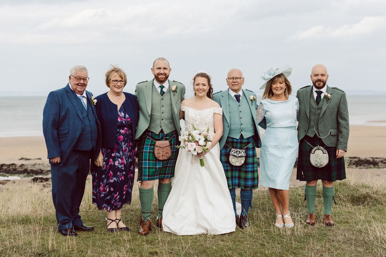 A group of wedding guests wearing traditional Scottish kilts and formal attire pose together on a grassy hillside by the ocean.