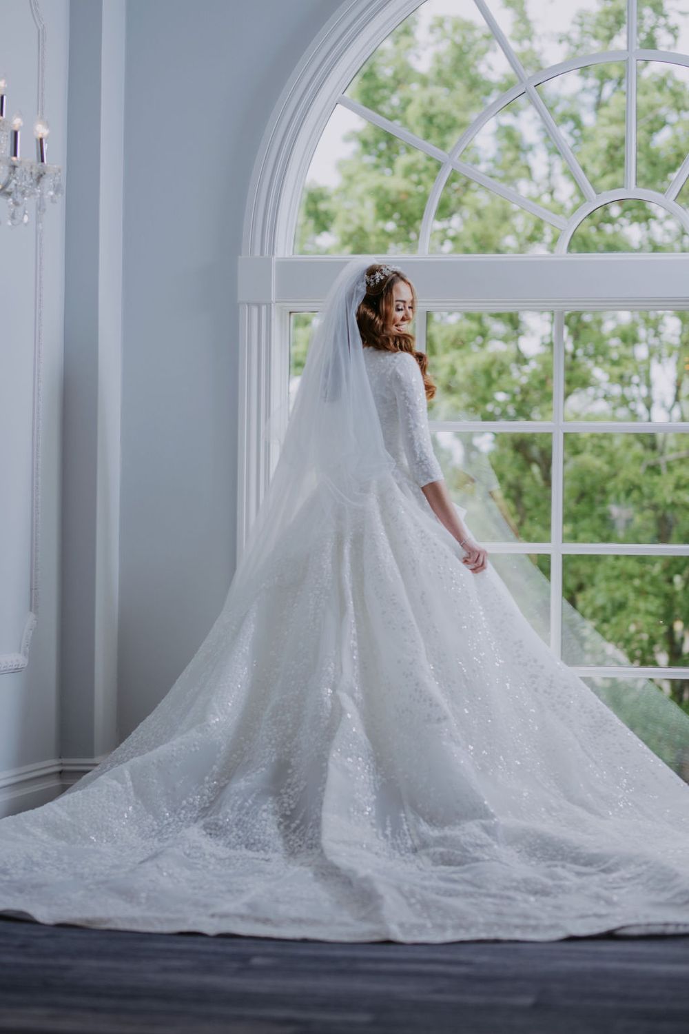 A bride in a luxurious white lace wedding gown with long train poses gracefully by sunlit windows in an elegant room.