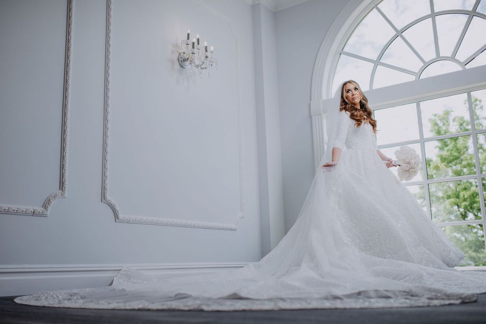 A bride in a flowing white wedding gown stands gracefully by an arched window in an elegant white room.