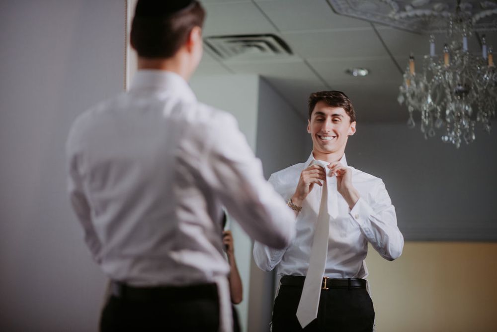 A person in a white dress shirt adjusts their tie while looking in a mirror with a chandelier visible in the background.