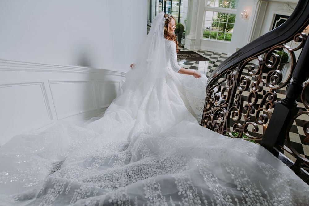 Flowing white wedding dress with intricate lace details cascades down an elegant staircase with ornate iron railings.