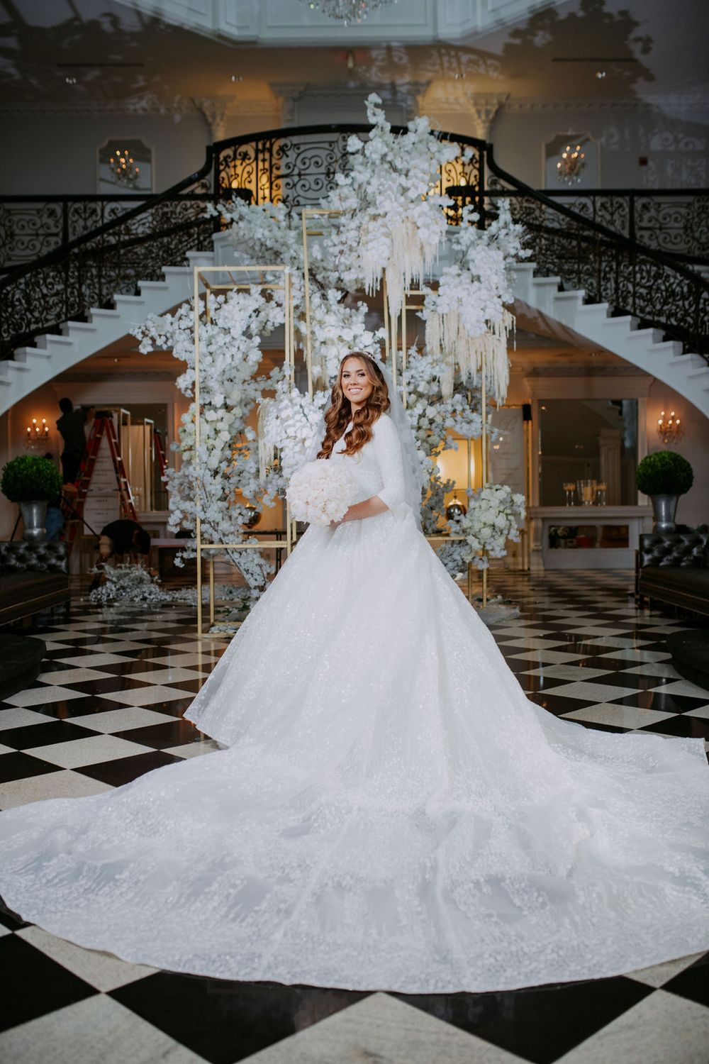 A stunning bridal portrait in a grand ballroom with checkered floors and an elegant white floral arch backdrop.